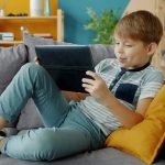 Băiat care stă pe canapea în casă și se uită pe tabletă