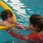 Fetiță care învață să înoate în piscină, ajutată de mama ei